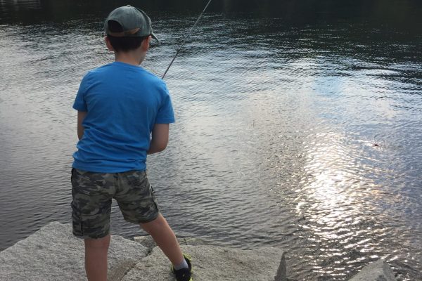 A boy in a blue shirt and camo shorts fishes from rocks by a lake surrounded by trees.