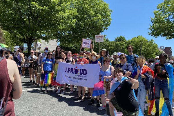 A group of people at a Pride parade hold a “J.Proud” banner, surrounded by rainbow flags and signs.