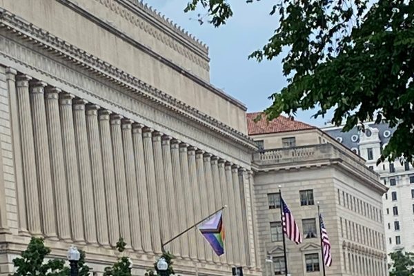 A building with columns, U.S. flags, and a Pride flag flying, with trees in the foreground.