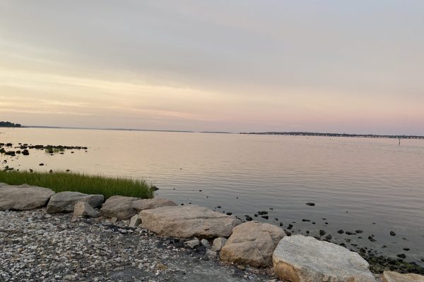 Rocky shoreline with calm water at sunset, sky glowing with soft pink and purple hues.