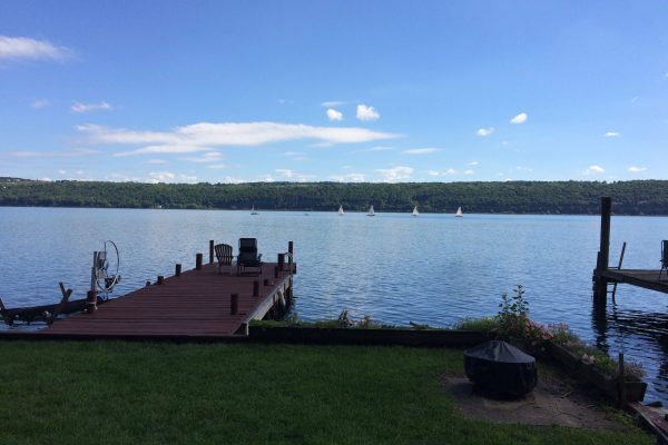 A wooden dock extends into a calm lake with sailboats in the distance and green hills beyond.
