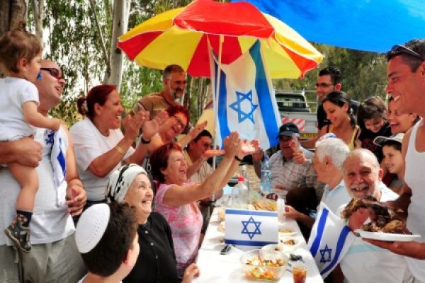 A lively outdoor gathering with people celebrating around a table, holding Israeli flags, and enjoying a meal.