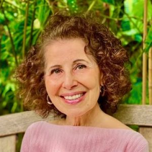 Smiling person with curly hair, wearing a pink top, seated outdoors with greenery in the background.