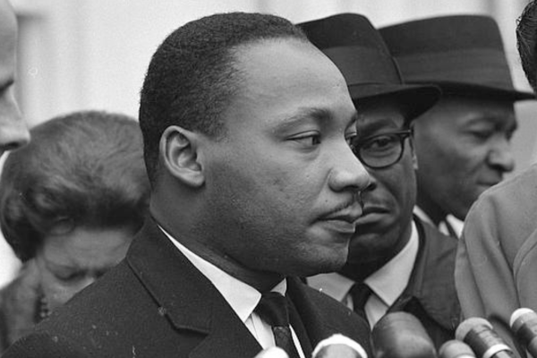 A man in a suit speaks into multiple microphones, surrounded by others outdoors.