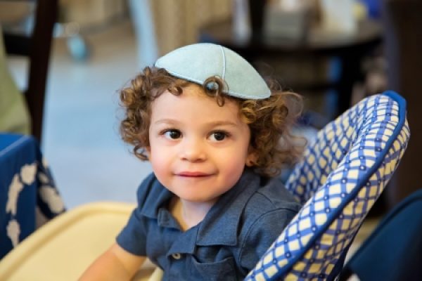 Curly-haired toddler wearing a blue kippah, sitting in a patterned high chair, smiling indoors.