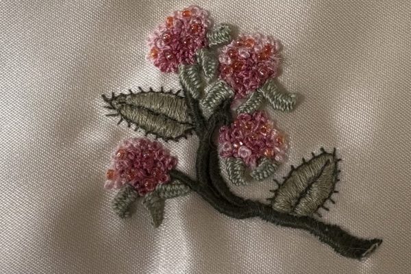 Embroidered branch with green leaves and clusters of pink flowers on a light fabric background.