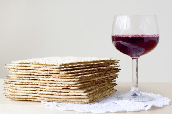 Stack of matzah next to a glass of red wine on a lace doily, against a plain background.