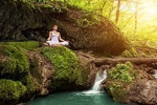 Person meditating on moss-covered rocks by a small waterfall in a sunlit forest.