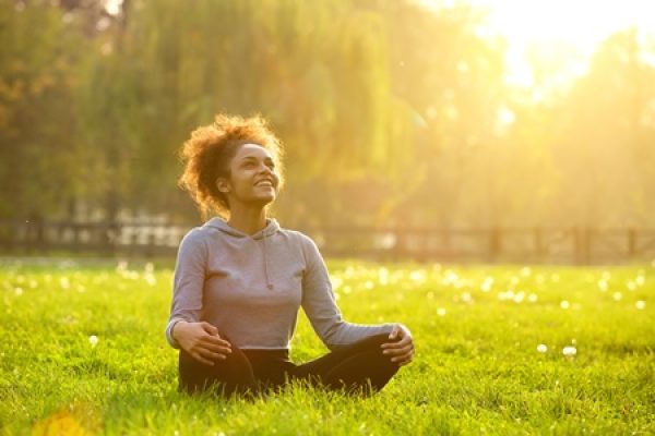 A person sitting cross-legged on grass, smiling and looking up, with sunlight filtering through the trees in the background.
