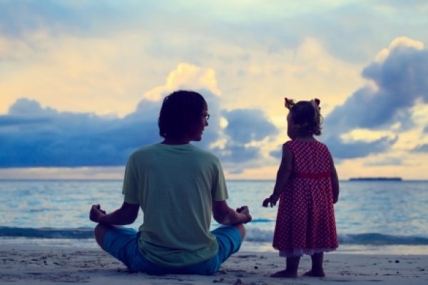 An adult and child sitting on a beach at sunset, facing the ocean.