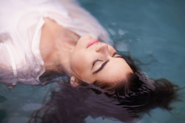 A woman in a white shirt floats calmly on water, with her eyes closed and hair submerged.
