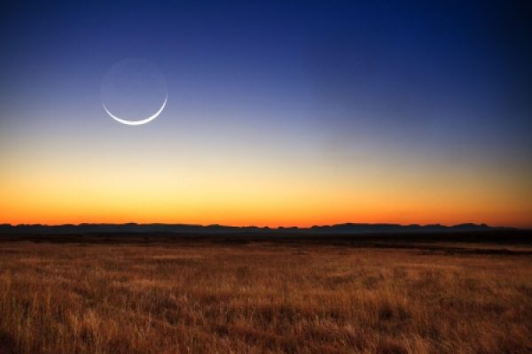 A crescent moon in a twilight sky over a vast, grassy plain with distant mountains on the horizon.