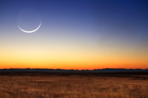 Crescent moon in a twilight sky over a vast grassy plain, with distant mountain silhouettes on the horizon.