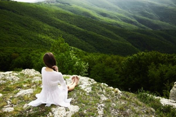 A woman in a white dress sits on a cliff edge, gazing at rolling green hills in the distance.