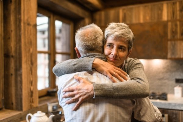 Older couple embracing warmly in a cozy kitchen with wooden furnishings and natural light.
