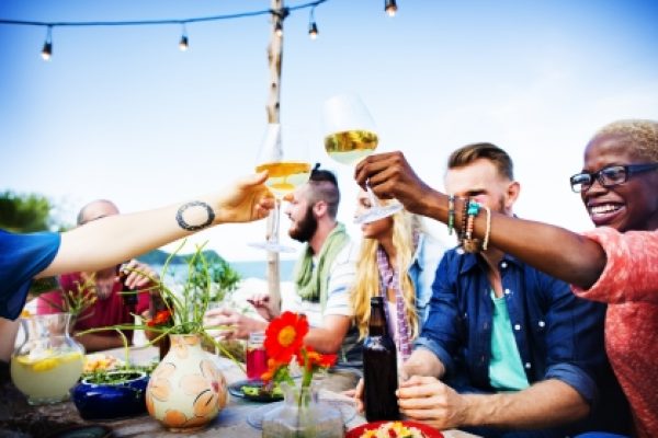 Group of people smiling and raising glasses in a toast at an outdoor gathering with food and drinks on the table.