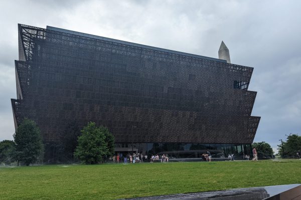 A modern, tiered museum building with a dark facade, trees, and people on the lawn; obelisk in background.
