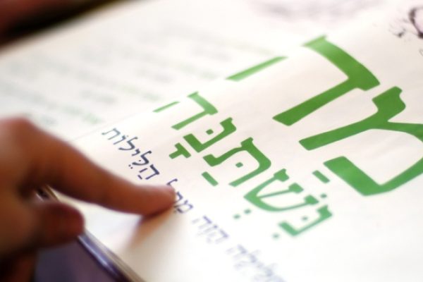 A finger points to Hebrew text in a book, focusing on large green letters.