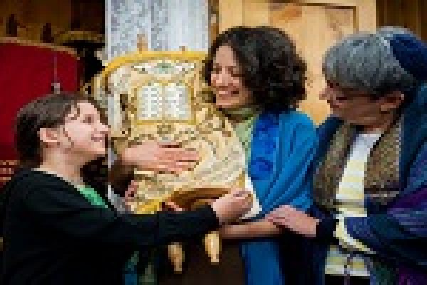 Three people smiling and holding a Torah scroll in a synagogue, with colorful curtains in the background.
