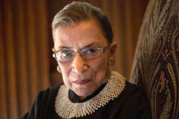 An older person wearing glasses and a lace collar, sitting on a patterned chair, with a serious expression.