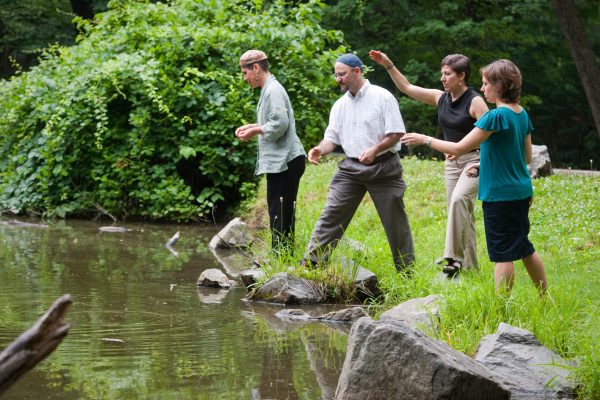 four people, some wearing kippot, at the edge of a lake throwing breadcrumbs to perform the tashlikh ritual