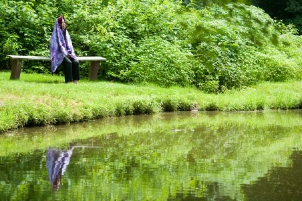 A person sits on a bench wearing a colorful scarf, gazing at a pond with lush greenery surrounding them.