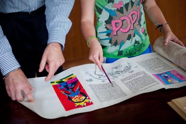 Two people point to illustrated and handwritten text on a scroll displayed on a table.