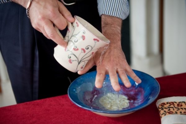 A person pours water from a decorated jug onto their hand over a blue bowl on a red tablecloth.