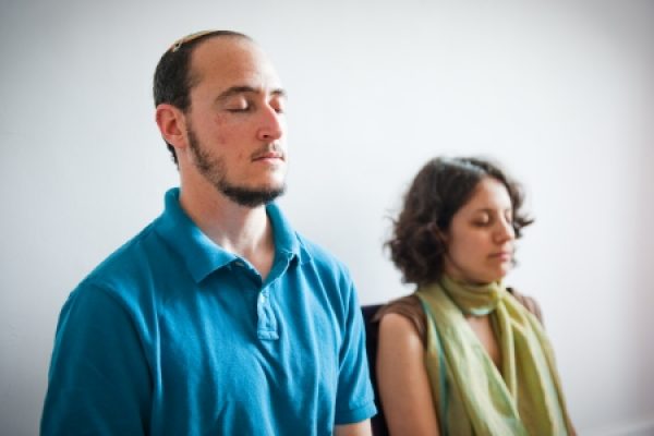 Two people sitting with eyes closed, appearing to meditate in a softly lit room.