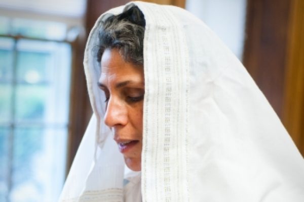 A person wearing a white headscarf, looking down, with a wooden window background.