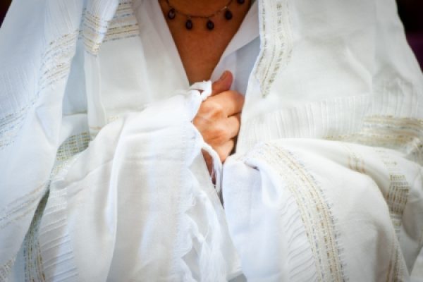 A person wearing a kittel and a white tallit and clasping the tallit at their chest