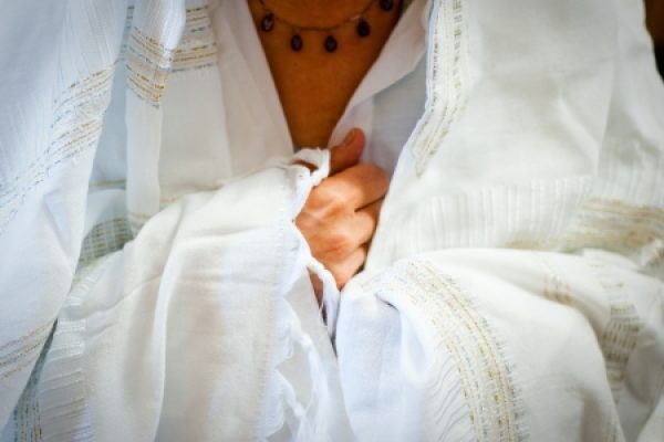 Person draped in a white prayer shawl, holding it close to their chest, wearing a beaded necklace.