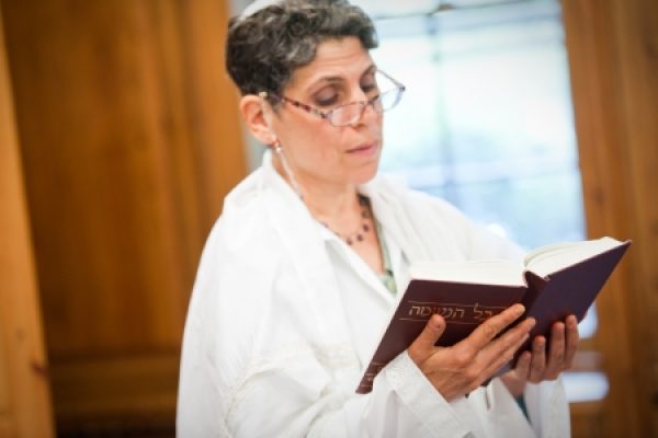Person in glasses reading a book, wearing a white garment in a softly lit room.