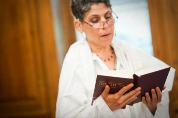 Person in a white garment reading a book with Hebrew text, standing indoors near a wooden surface.