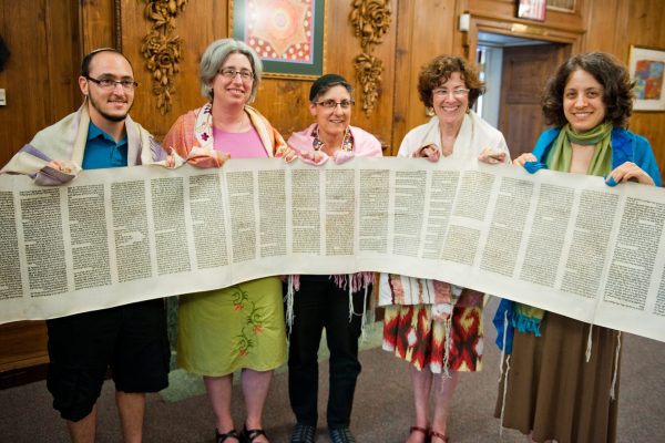 Five people smiling and holding an open Torah scroll in a wood-paneled room.