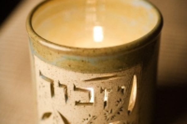 A lit memorial candle in a ceramic holder with Hebrew letters on a table.
