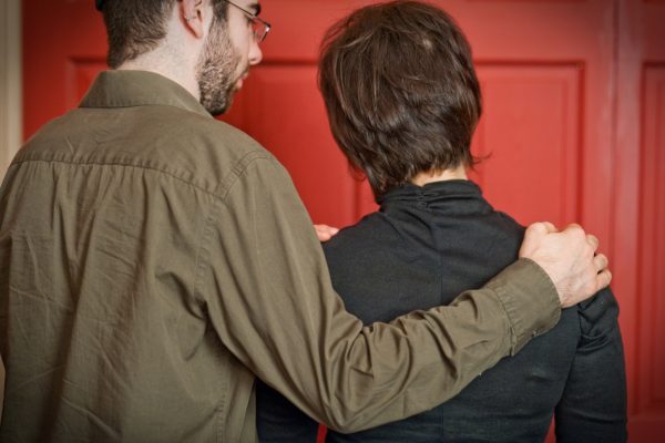 A man with glasses embraces a woman in front of a red door, both facing away from the camera.