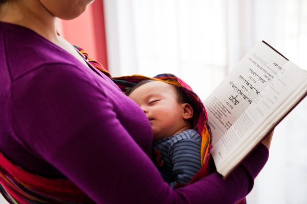 A woman reads a book in Hebrew while holding a sleeping baby in a sling.