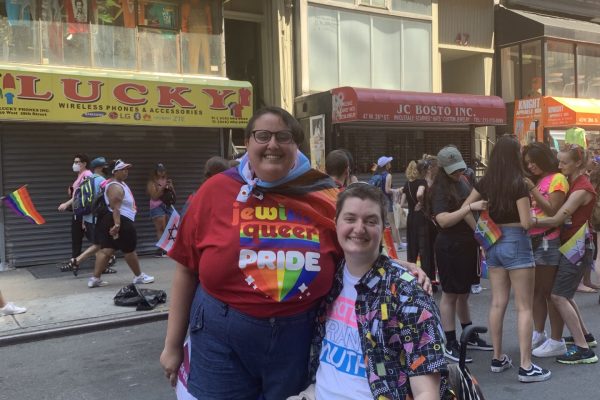 Two people at a pride parade, one standing with a rainbow flag, the other in a wheelchair, both smiling.