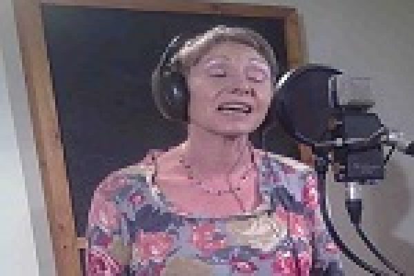 A woman sings into a microphone in a recording studio wearing headphones and a floral top.