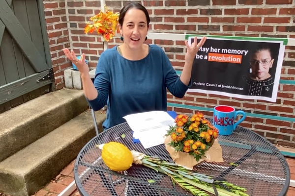 A woman smiling at a table with flowers, a lemon, a blue mug, and a poster on the brick wall behind her.