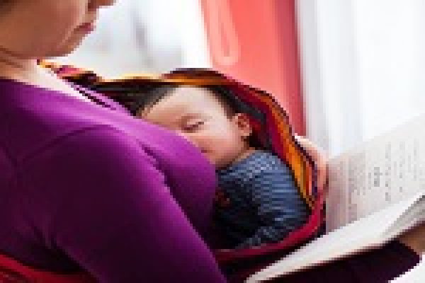 A person holding a sleeping baby in a sling while reading a book.