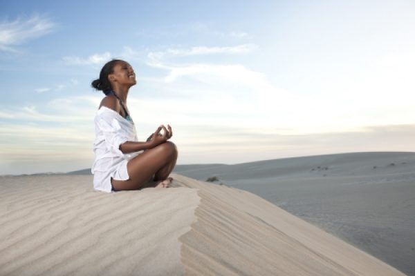 Person meditating on a sandy dune under a clear sky, wearing a white outfit, facing the sunrise or sunset.