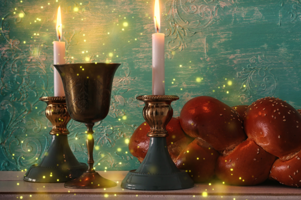 Two lit candles, a goblet, and challah bread on a table with a teal background and glowing lights.