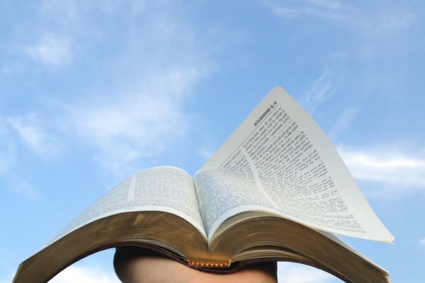 A hand holds an open book up to a blue sky with scattered clouds.