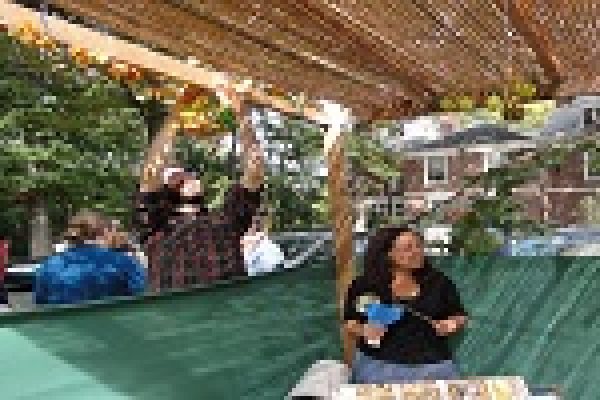 People decorating a sukkah with leaves; a woman sits inside smiling.