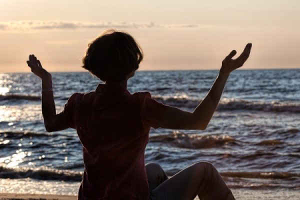 Person sitting on a beach at sunset, arms raised, facing the ocean waves.