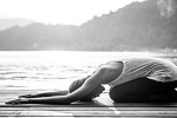Person in childs pose on a wooden deck by a lake, with mountains in the background. Black and white image.