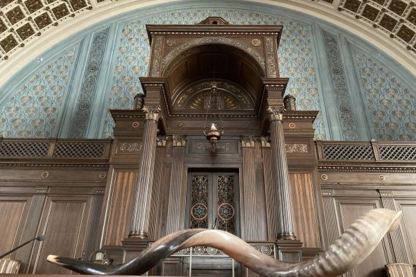 A curved shofar rests on a wooden surface in front of an ornate synagogue ark and blue mural.