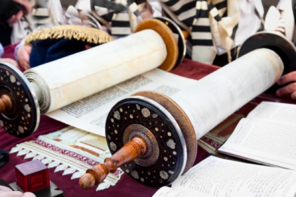 Two Torah scrolls on a table with open books and people nearby, wearing traditional garments.